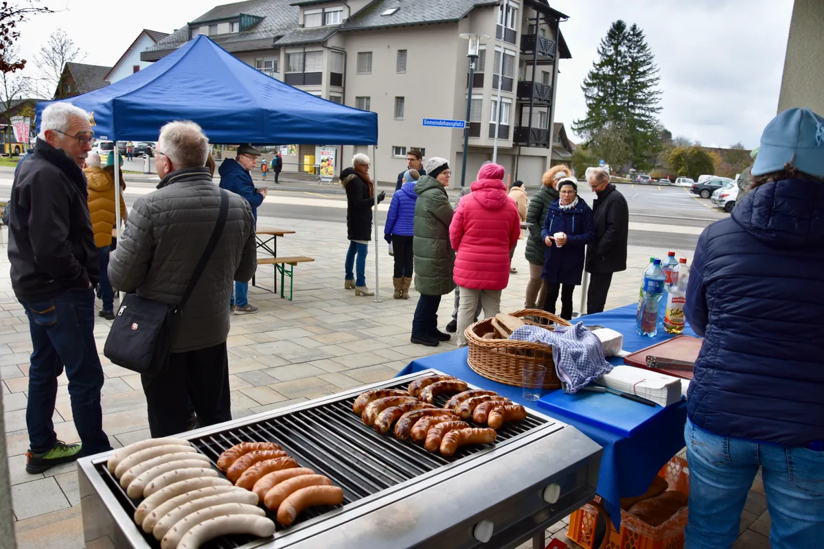 Bei frostigen Temperaturen gab es nebst Diskussionen über das neue Dorfzentrum auch noch Würste serviert. Menschen stehen auf einem Platz. Auch ein Grill mit Würsten ist zu sehen.